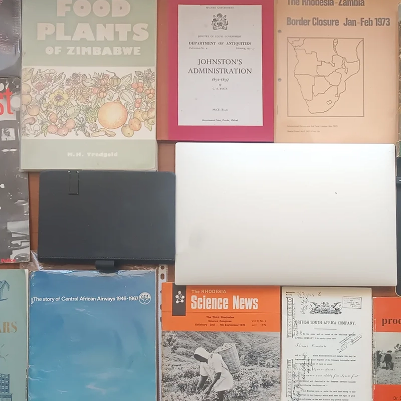 Archival materials laid out on a work surface at Abantu Archives, Harare: includes Food Plants of Zimbabwe (Tredgold), Johnston's Administration 1891–1897 (Department of Antiquities, Malawi), The Rhodesia–Zambia Border Closure Jan–Feb 1973, The Matopos, Outpost magazine, Reflections of a Son of the Soil, The Story of Central African Airways 1946–1967, Rhodesia Science News, a British South Africa Company transfer document, Cattle Production, and Miracle Valley (Frank Muss).